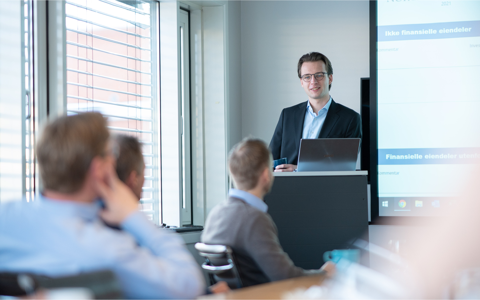 En mann med briller og moerkt haar staar foran en skjerm og snakker til flere mennesker som hoerer paa.